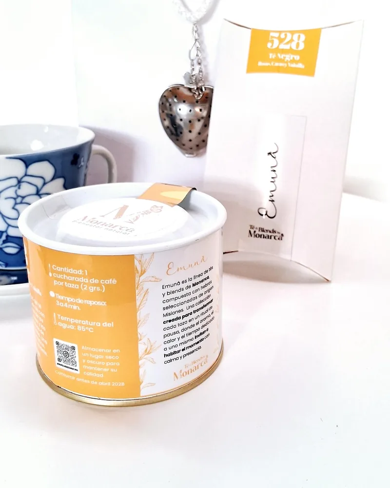 Lata de té Emuná 528 con sobre y filtro en forma de corazón, junto a una taza azul y blanca